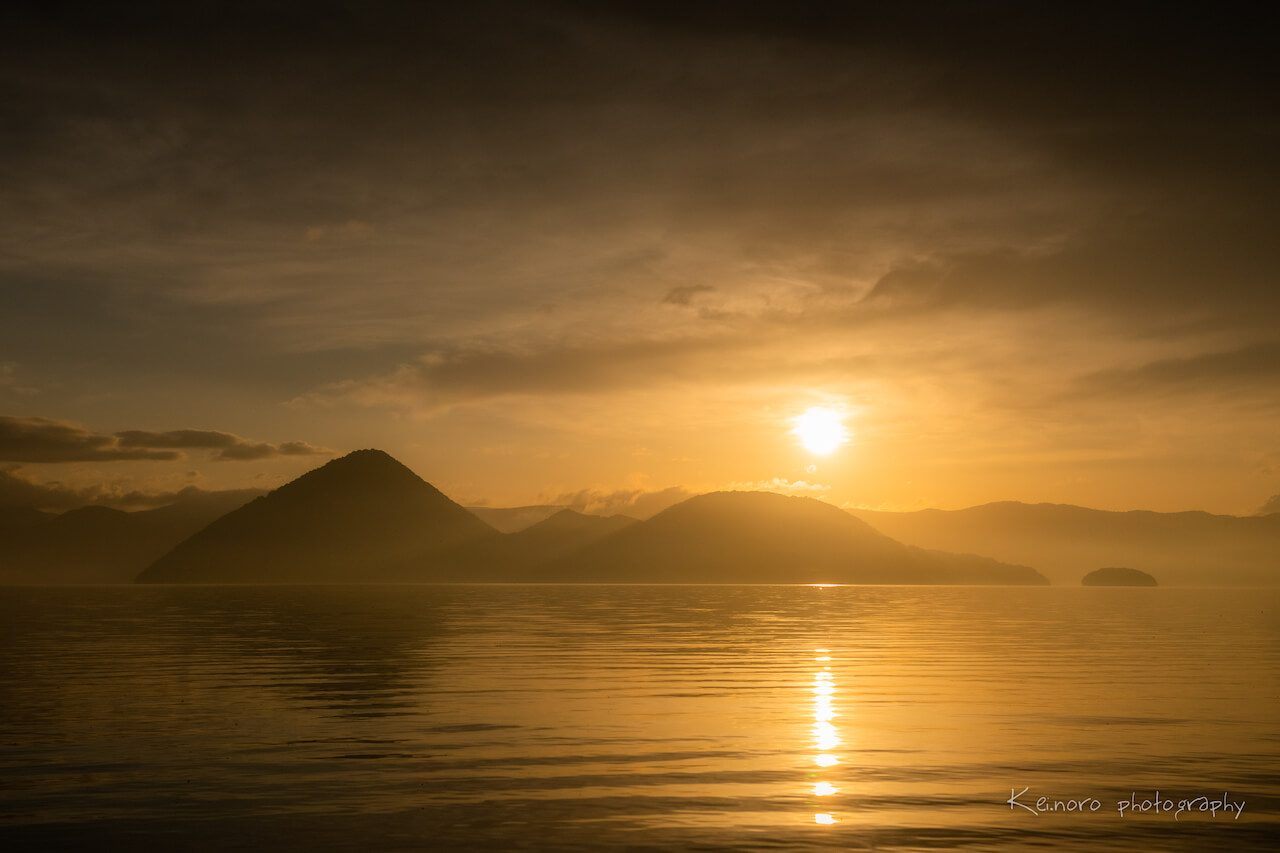 The sunrise over Lake Toya, captured from Tsukiura early in the morning, in May. The water is particularly transparent from winter to spring, and the light of the morning sun is beautifully reflected on the surface of the lake on days when the waves are calm. ©Noro Keiichi